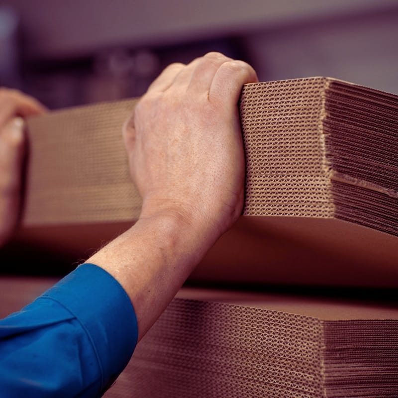 Krabice a obaly na míru: jak významně snížit logistické náklady? 25 Detailní záběr na ruce držící vrstvy vlnité lepenky, připravené k dalšímu zpracování nebo výrobě obalových materiálů